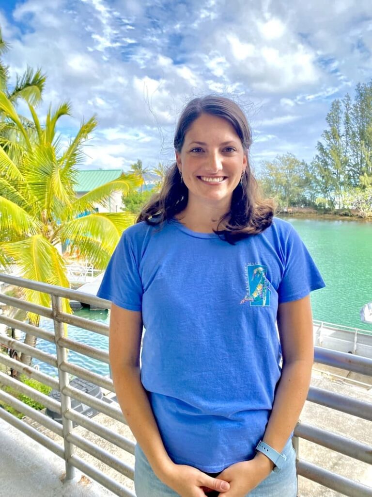 Woman smiling on a balcony by water, with palm trees and a bright, partly cloudy sky in the background.