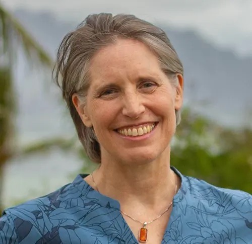 Smiling woman with short gray hair, wearing a blue patterned shirt and pendant, outdoors with greenery.
