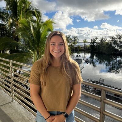 Smiling woman in a brown shirt stands by a railing with palm trees, water, and boats in the background.