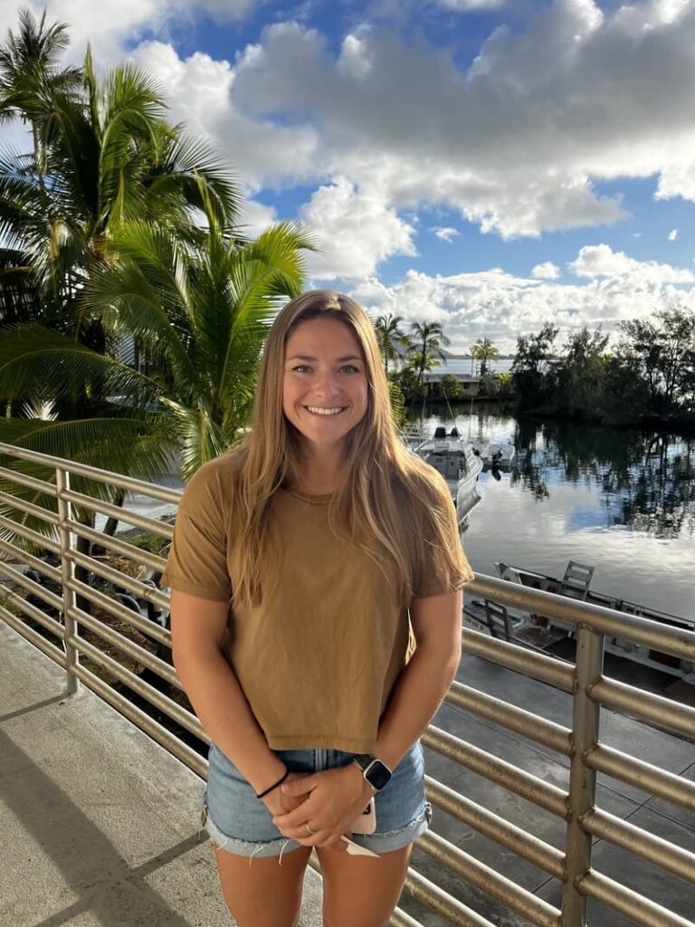 Smiling woman in a brown shirt stands by a railing with palm trees, water, and boats in the background.