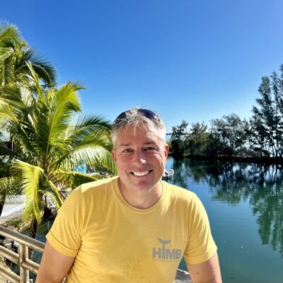Smiling man in a yellow shirt by water, with palm trees and blue sky in the background.