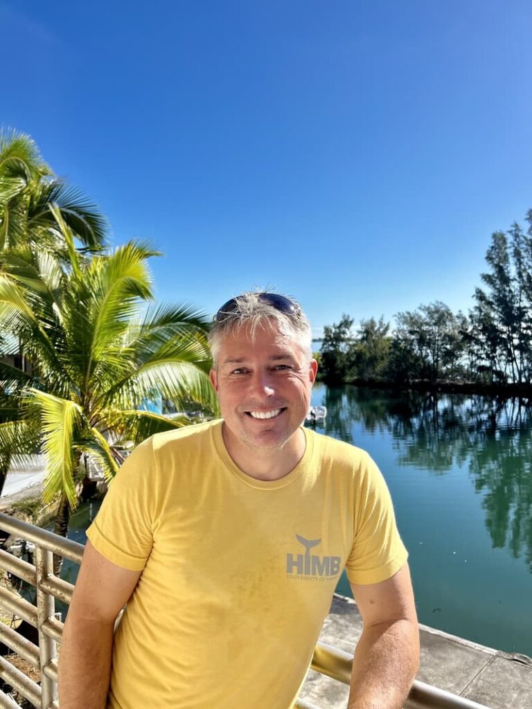 Smiling man in a yellow shirt by water, with palm trees and blue sky in the background.
