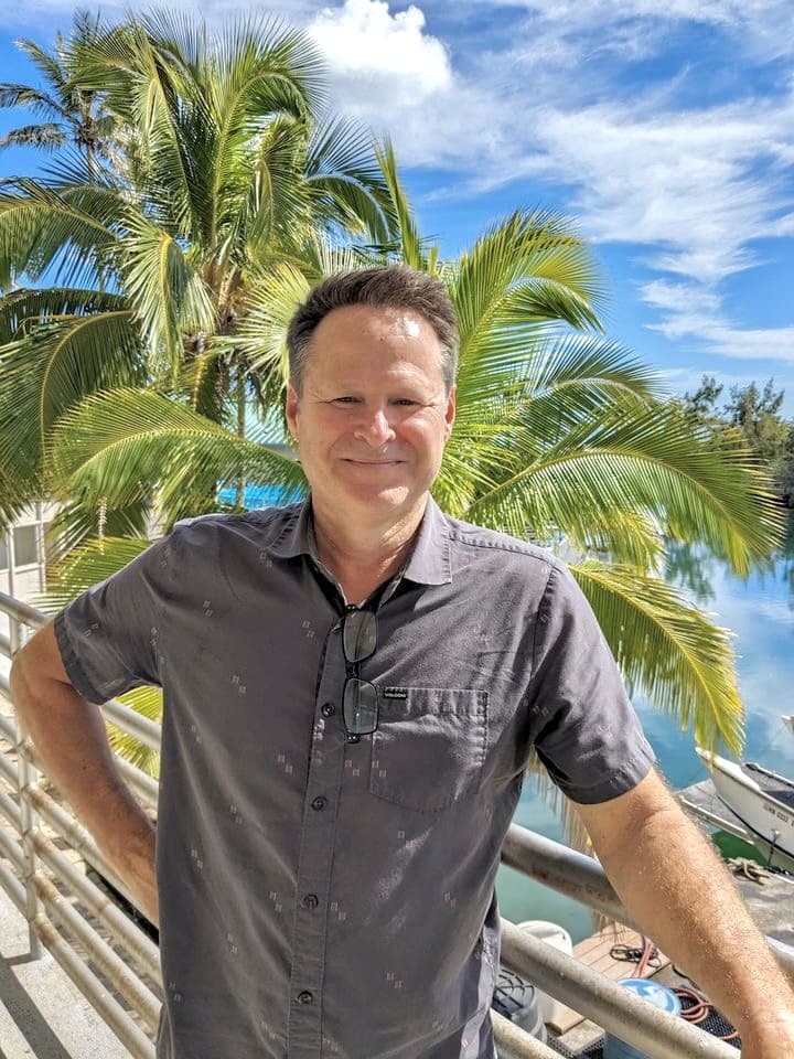 Smiling man in a gray shirt standing by a railing with palm trees and water in the background.