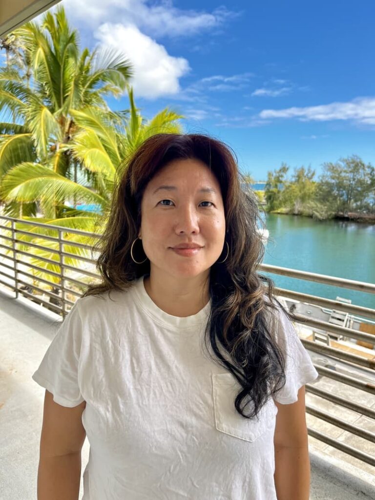 Woman in a white t-shirt standing on a balcony by water, with palm trees and blue sky in the background.