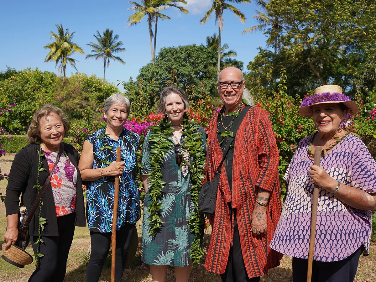 Five smiling people in colorful clothing and leis stand outdoors with trees and flowers in the background.