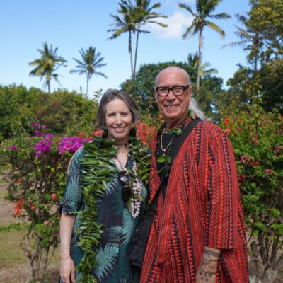 A smiling man and woman in tropical attire stand together outdoors with palm trees and flowers behind them.