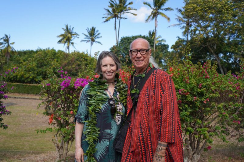 A smiling man and woman in tropical attire stand together outdoors with palm trees and flowers behind them.