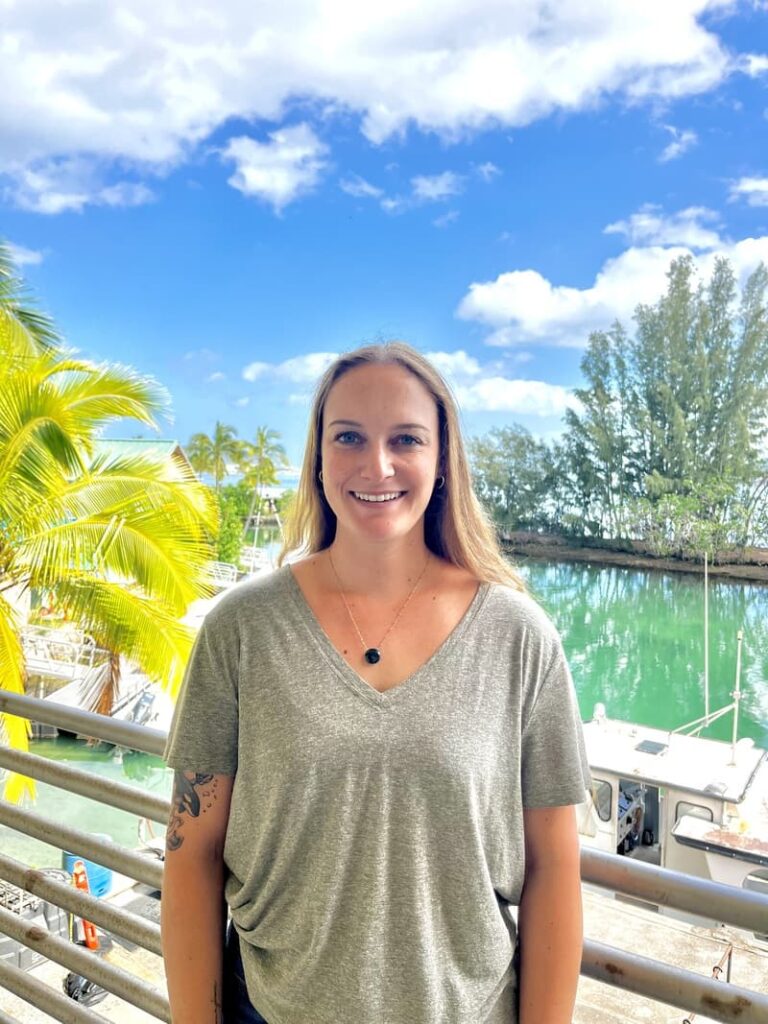 Woman smiling on a balcony near water, lush trees, boats, and a bright blue sky with scattered clouds.