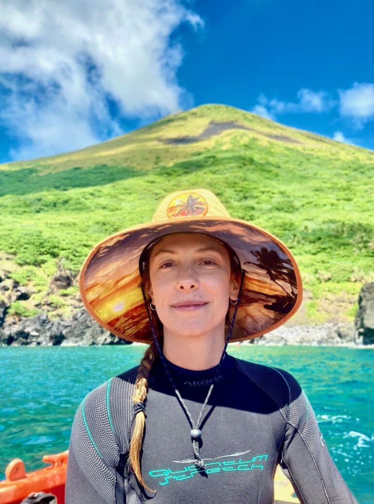 A woman in a sunhat and wetsuit smiles by turquoise water with a lush green mountain in the background.