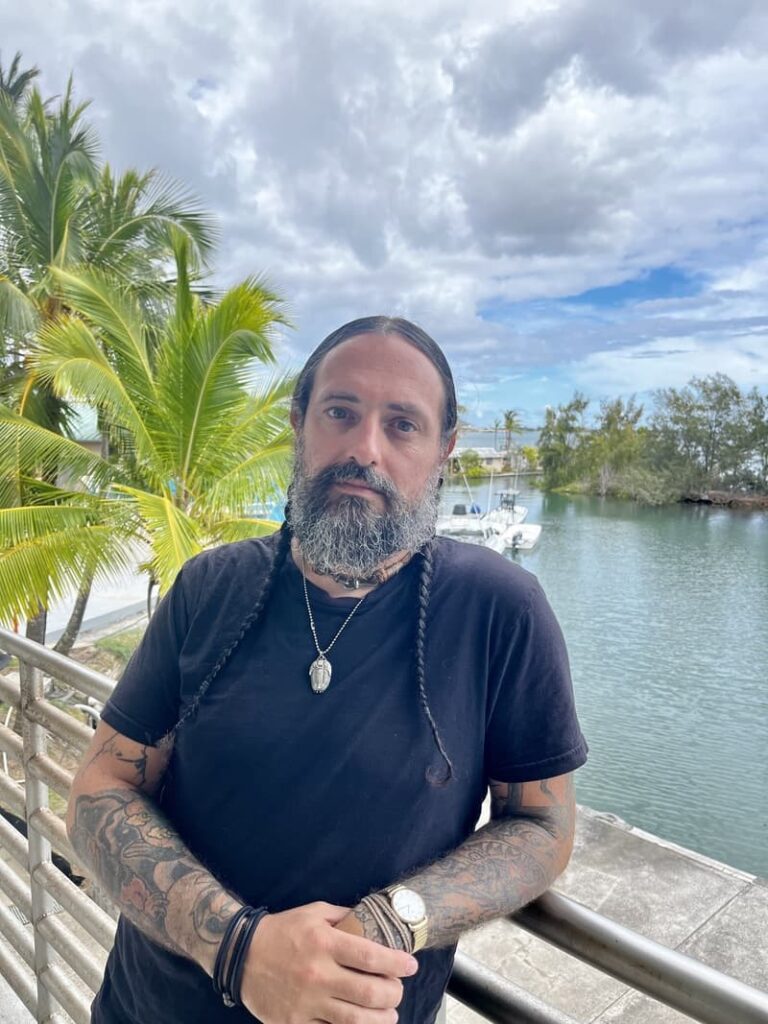 A bearded man with braided hair stands by a waterfront railing, palm trees and a boat in the background.