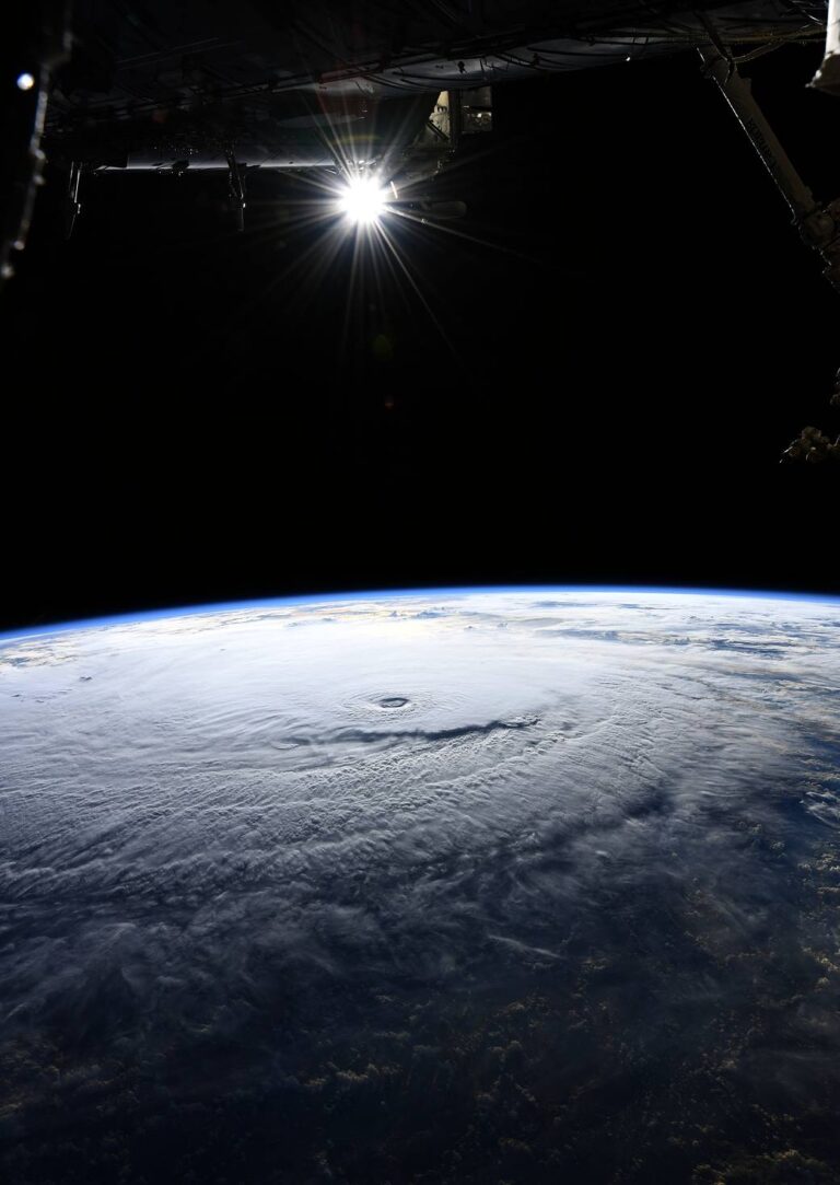 View of a hurricane’s swirling clouds on Earth from space, with part of a spacecraft and the sun visible above.