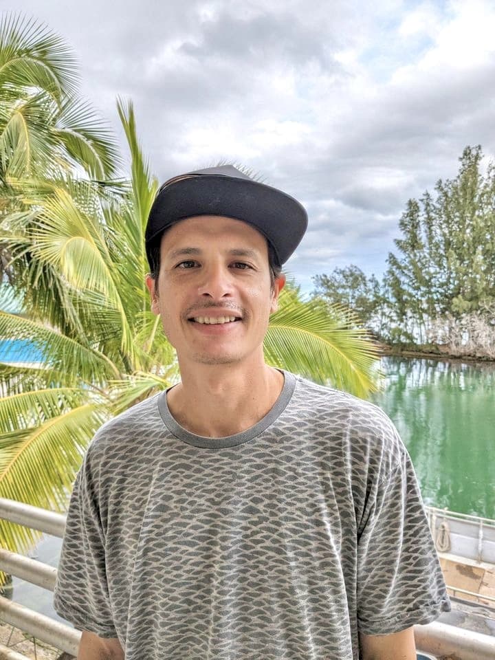 A man in a cap smiles outdoors near palm trees and water under a cloudy sky.