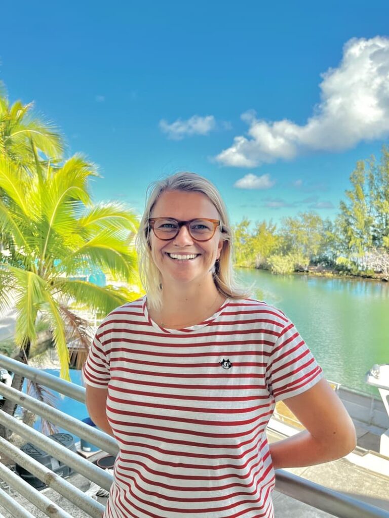 Smiling woman in glasses and striped shirt stands by water, with palm trees and a blue sky in the background.