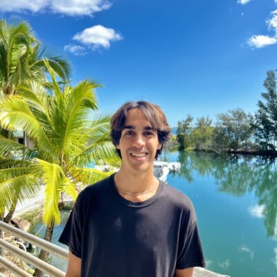 A young man in a black shirt stands by water and palm trees under a bright blue sky with clouds.