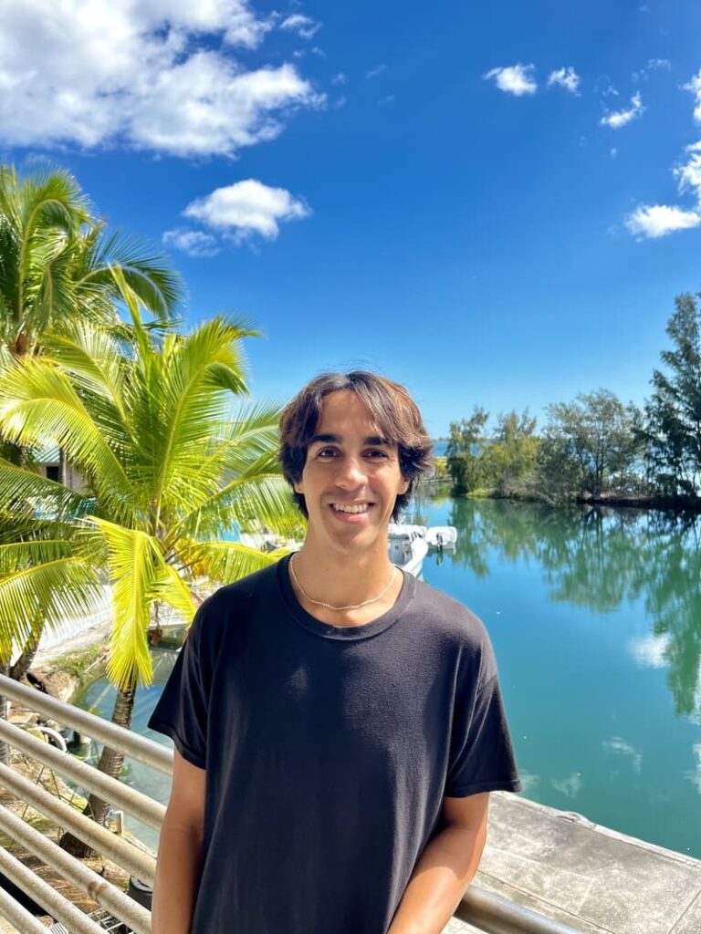 A young man in a black shirt stands by water and palm trees under a bright blue sky with clouds.