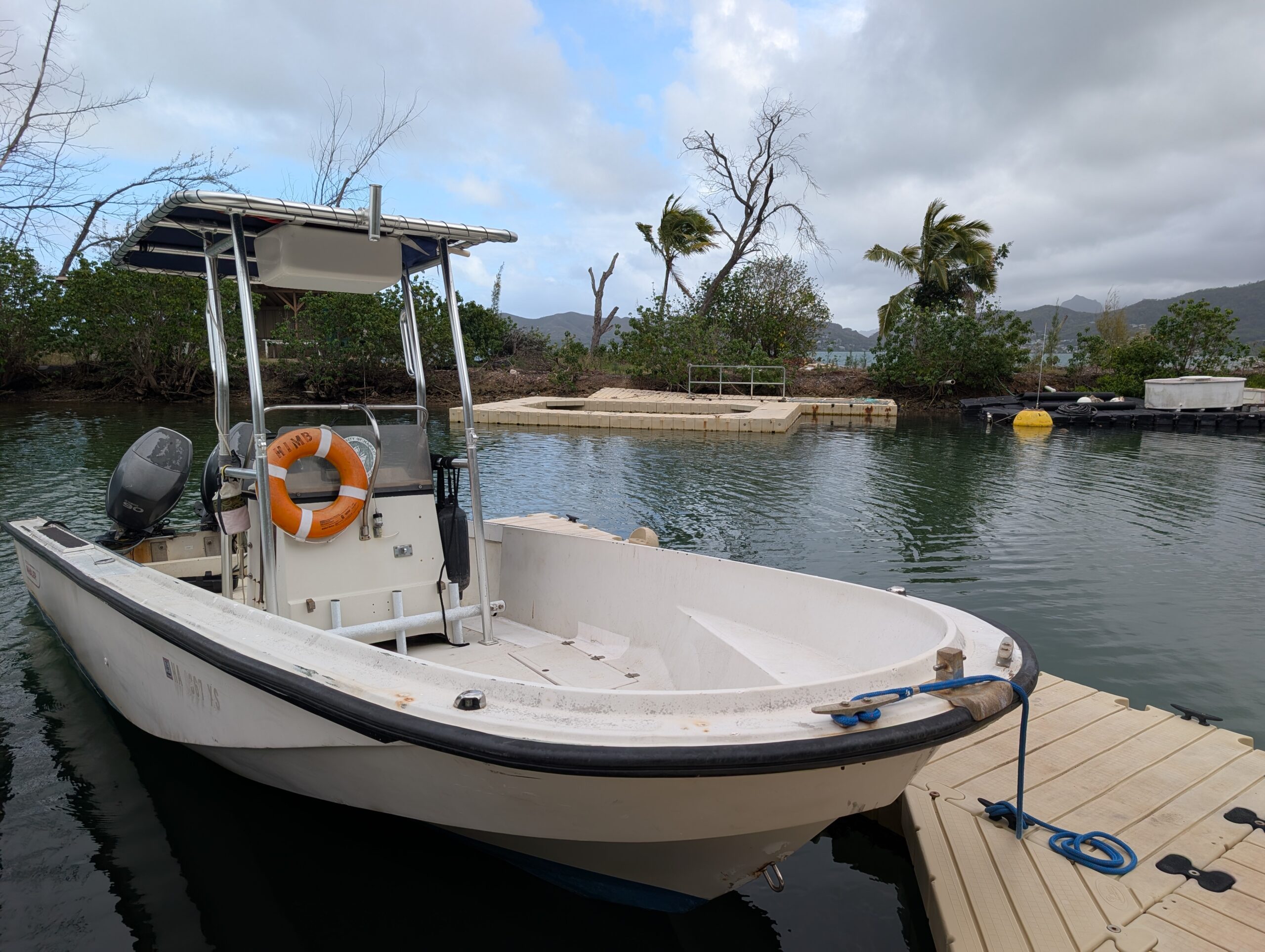 A small white motorboat is docked on a floating pier in a calm, tropical inlet with trees and cloudy skies.