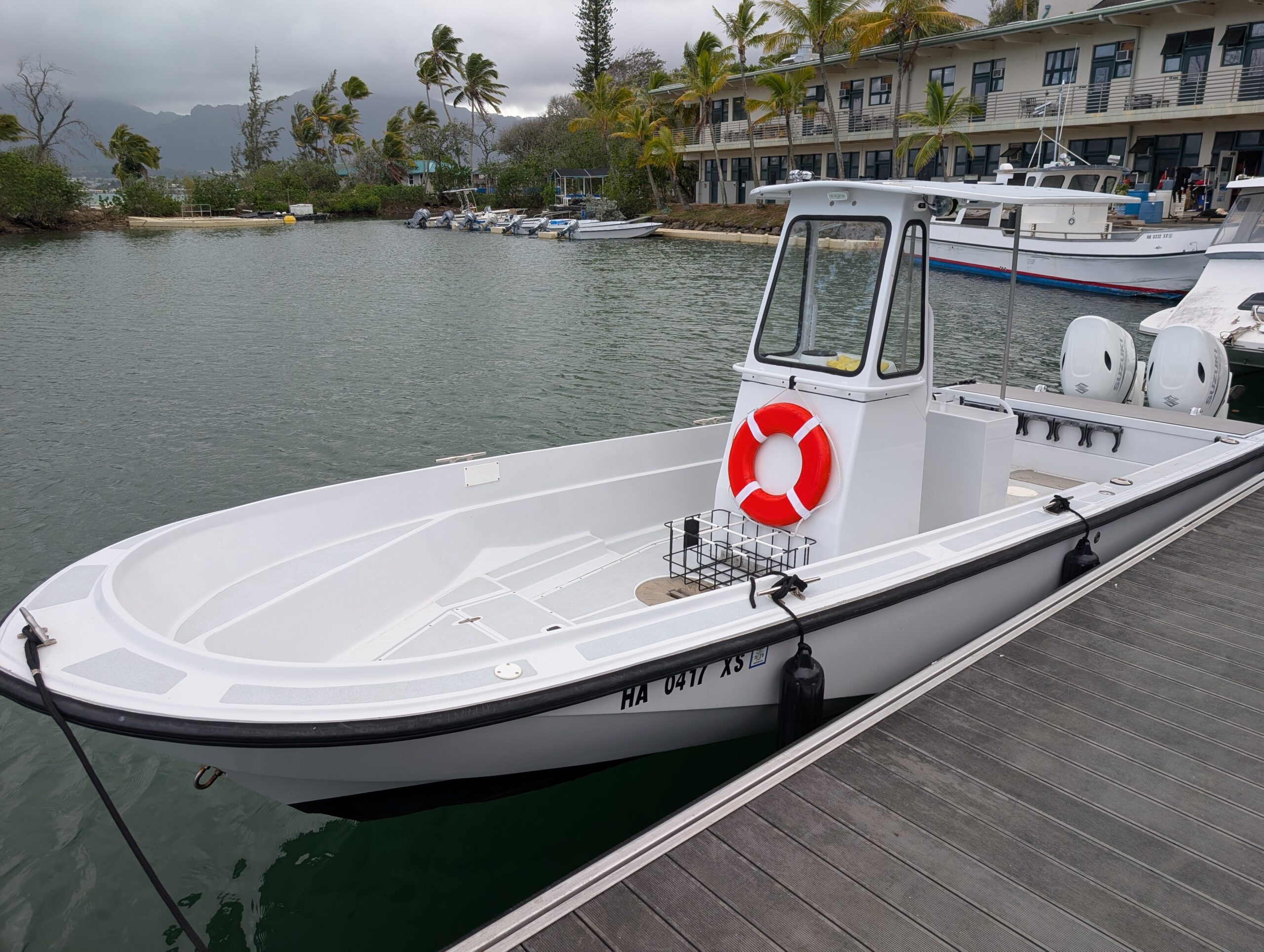 A white boat with a red life preserver is docked by the water near palm trees and a building.