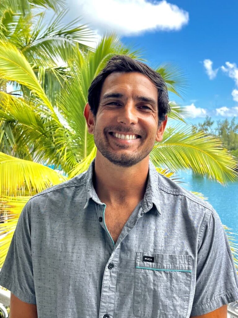 Man smiling outdoors with palm trees and blue water in the background under a bright, sunny sky.