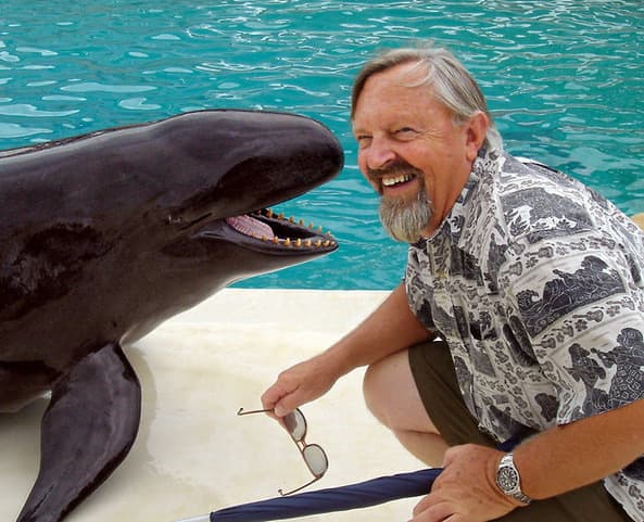 Smiling man kneels beside a black dolphin-like marine animal near a pool, holding his glasses.