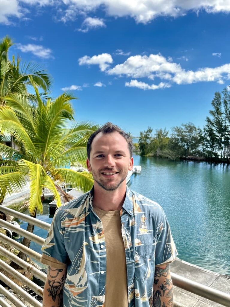 Smiling man in a tropical shirt stands by water with palm trees and blue sky in the background.