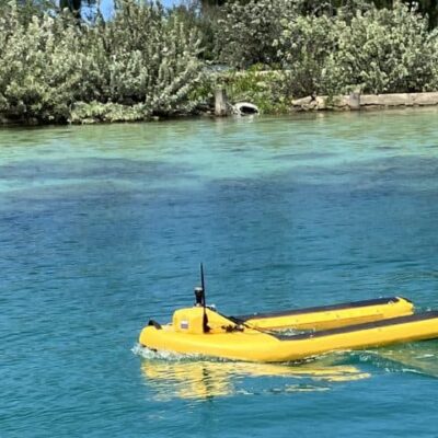 A small yellow robotic boat floats on clear blue water near a shoreline with dense green bushes.