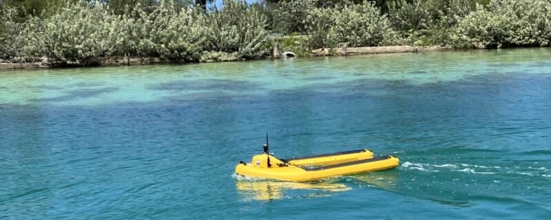 A small yellow robotic boat floats on clear blue water near a shoreline with dense green bushes.