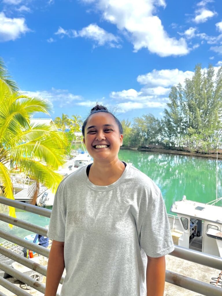 Smiling person in a gray shirt stands by a marina with boats, palm trees, and blue sky in the background.