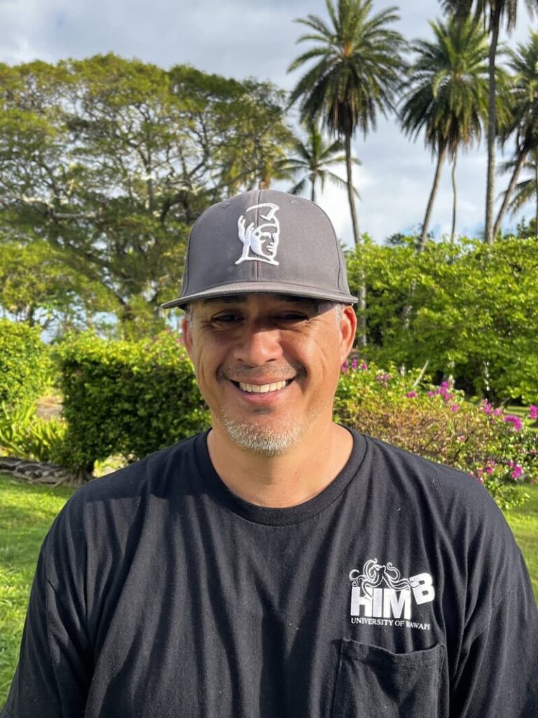 Smiling man in a black University of Hawaii HMB shirt and hat stands outdoors with palm trees and greenery behind him.