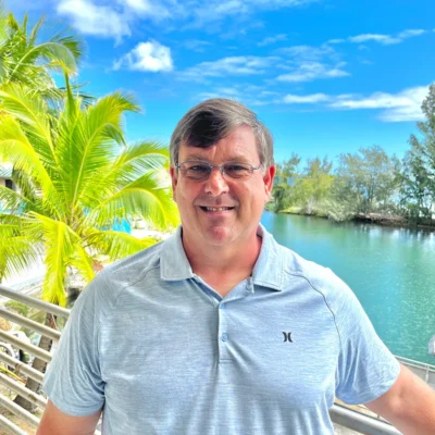 Smiling man in glasses stands by water with palm trees and blue sky in the background.