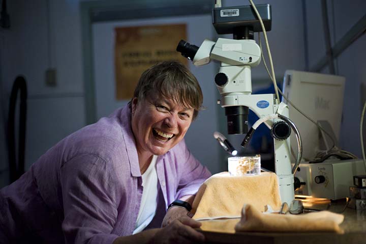 Person smiling beside a microscope in a lab, wearing a light purple shirt, with lab equipment visible.