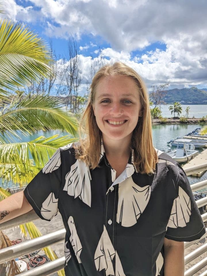 Smiling person in a ghost-patterned shirt stands by a railing with palm trees, water, and mountains behind.