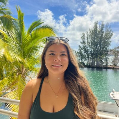 Woman with long hair and sunglasses poses by tropical water with palm trees and blue sky in the background.