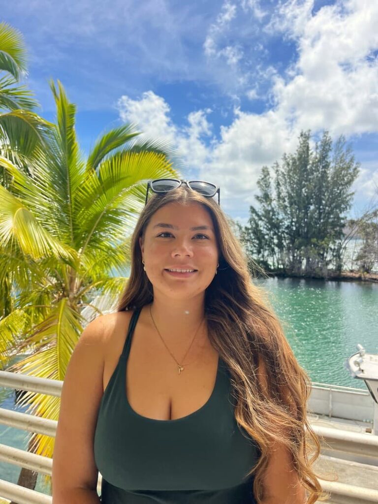 Woman with long hair and sunglasses poses by tropical water with palm trees and blue sky in the background.