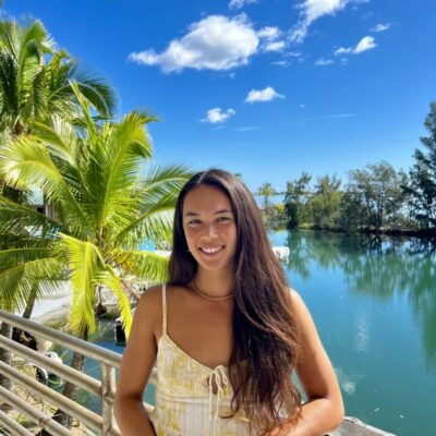 Woman with long brown hair smiles by a scenic lake, palm trees, and blue sky with clouds in the background.
