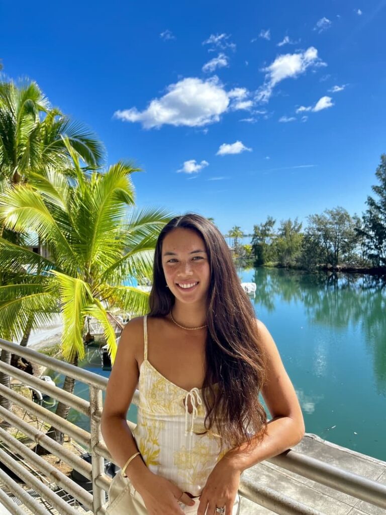 Woman with long brown hair smiles by a scenic lake, palm trees, and blue sky with clouds in the background.