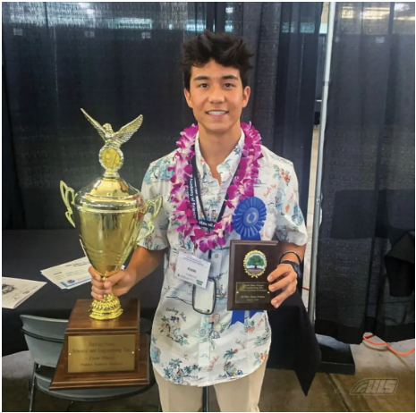 Smiling young man in a floral shirt holding a trophy, a plaque, and wearing a lei and a blue ribbon.
