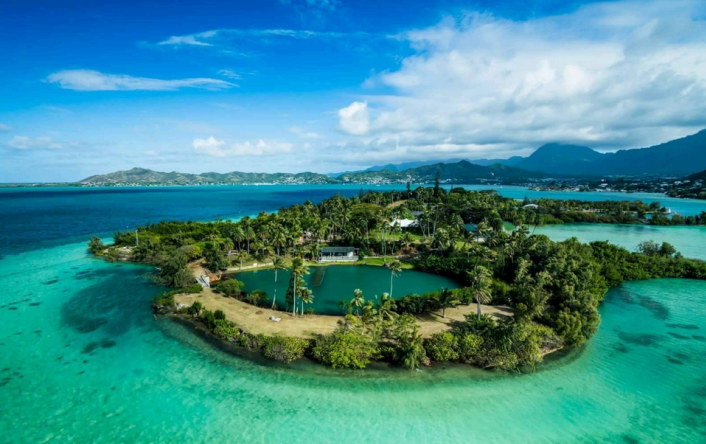 A lush, green island surrounded by clear turquoise waters under a bright blue sky with distant mountains.