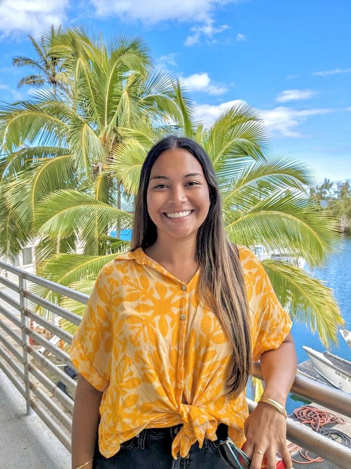 Smiling woman in a yellow shirt stands by a railing with palm trees and water in the background.