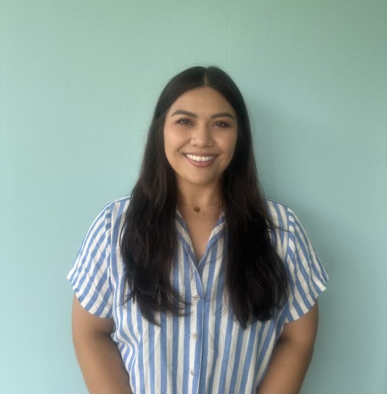 A woman with long dark hair smiles, standing against a light blue background, wearing a blue striped shirt.