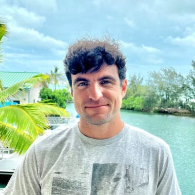 Man in a gray T-shirt standing outside near water, with trees and a boat in the background on a cloudy day.