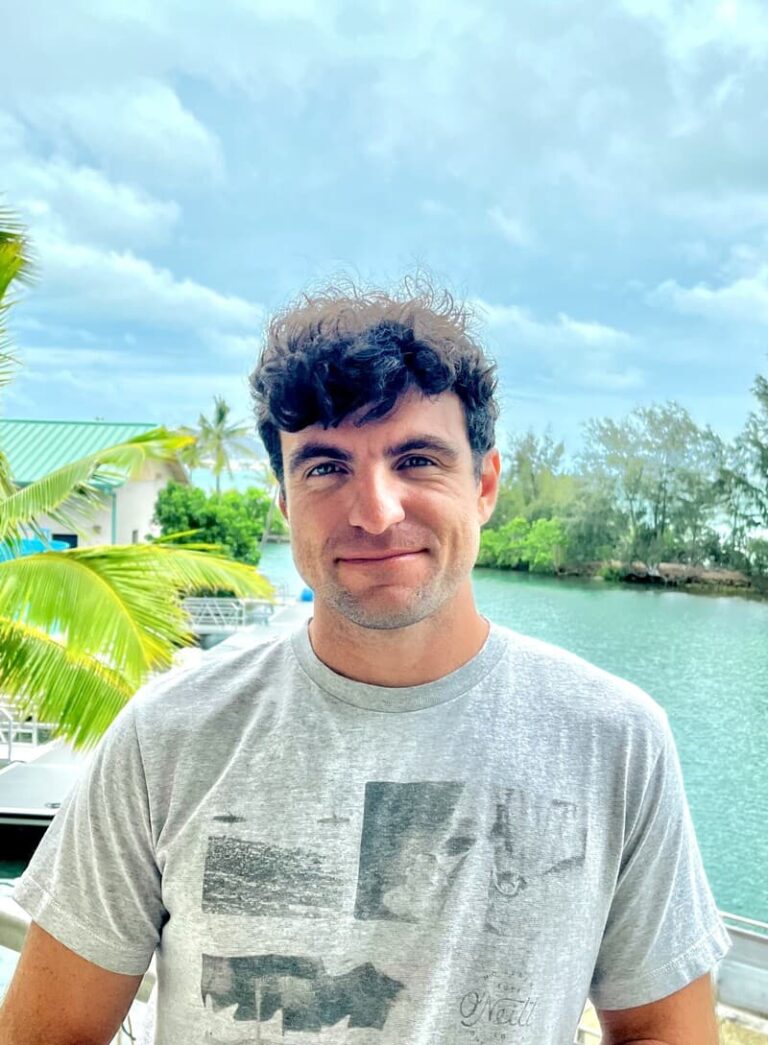 Man in a gray T-shirt standing outside near water, with trees and a boat in the background on a cloudy day.