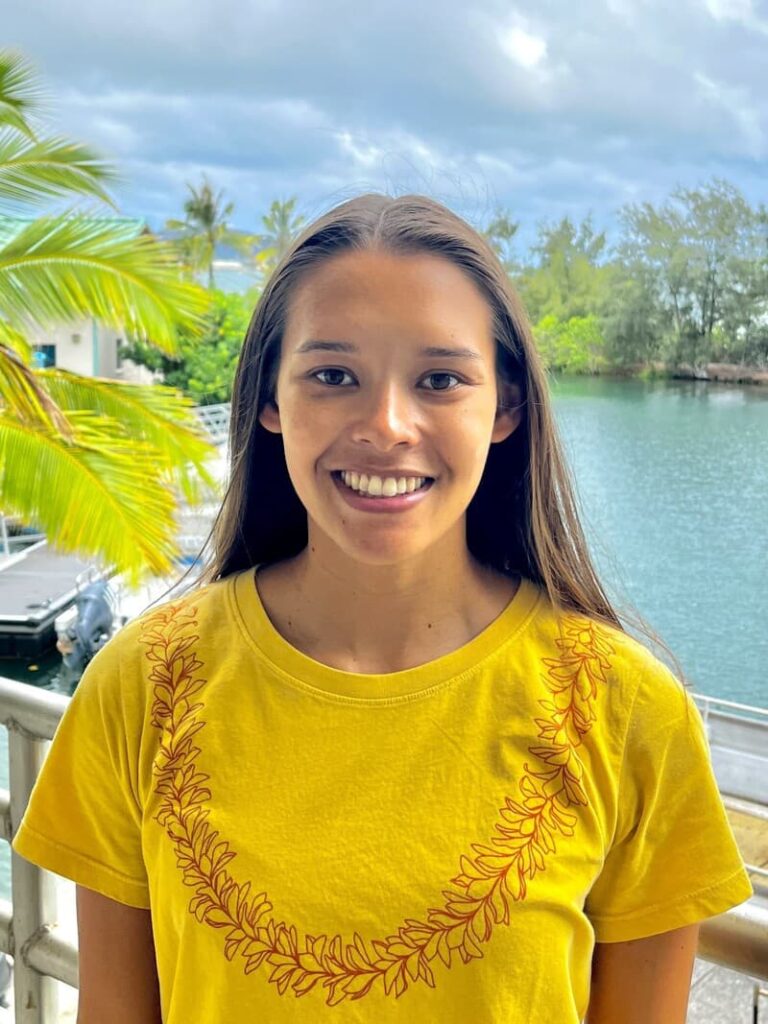 Young woman in a yellow shirt smiles outdoors near water, with palm trees and cloudy sky in the background.