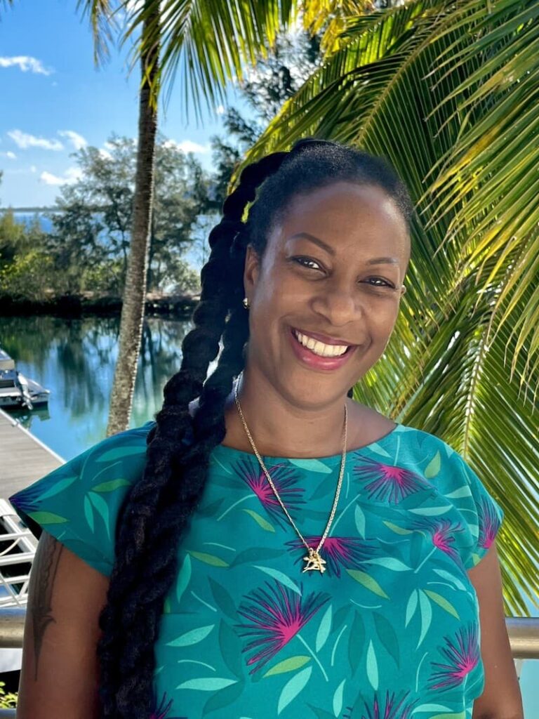 Smiling woman with long braids, wearing a colorful dress, stands by palm trees near water on a sunny day.