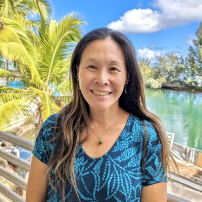 Woman smiling outdoors by water with palm trees, wearing a blue patterned shirt under a bright blue sky.