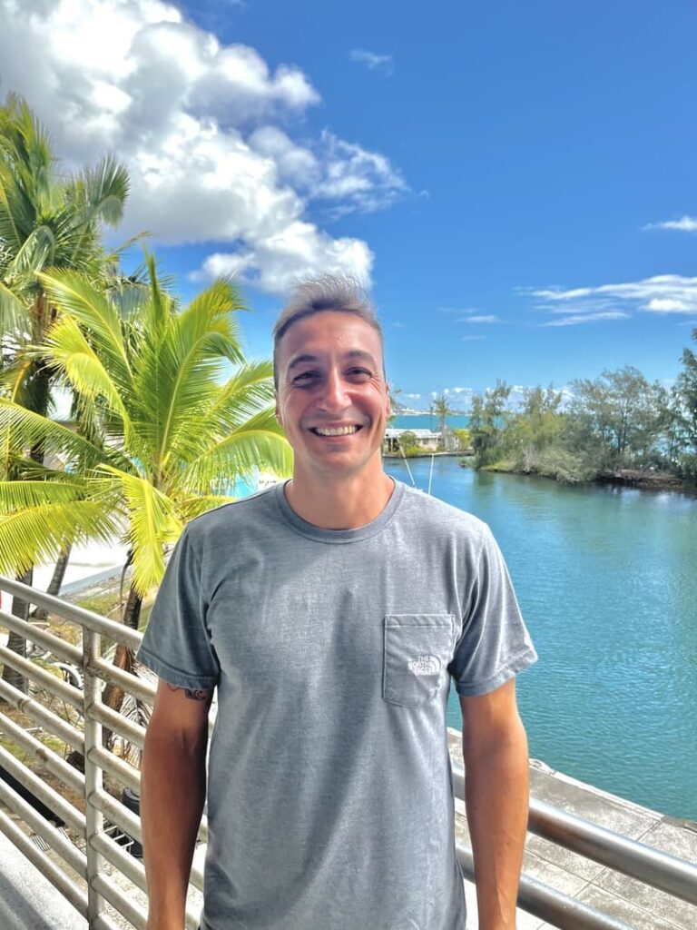 Man smiling on a balcony with palm trees, blue sky, and water in the background.
