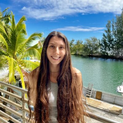 A woman with long brown hair smiles by a railing near water, palm trees, and a partly cloudy sky.