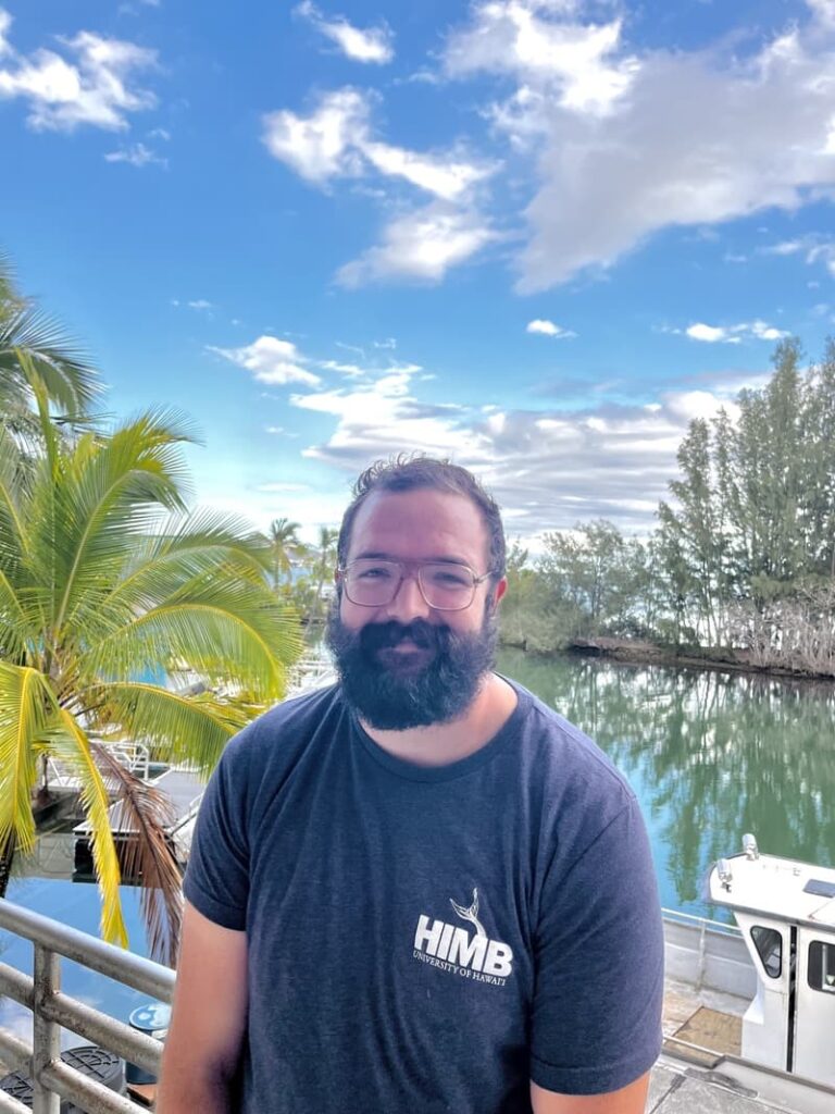 A man with a beard and glasses stands by water with boats, palm trees, and a blue sky in the background.