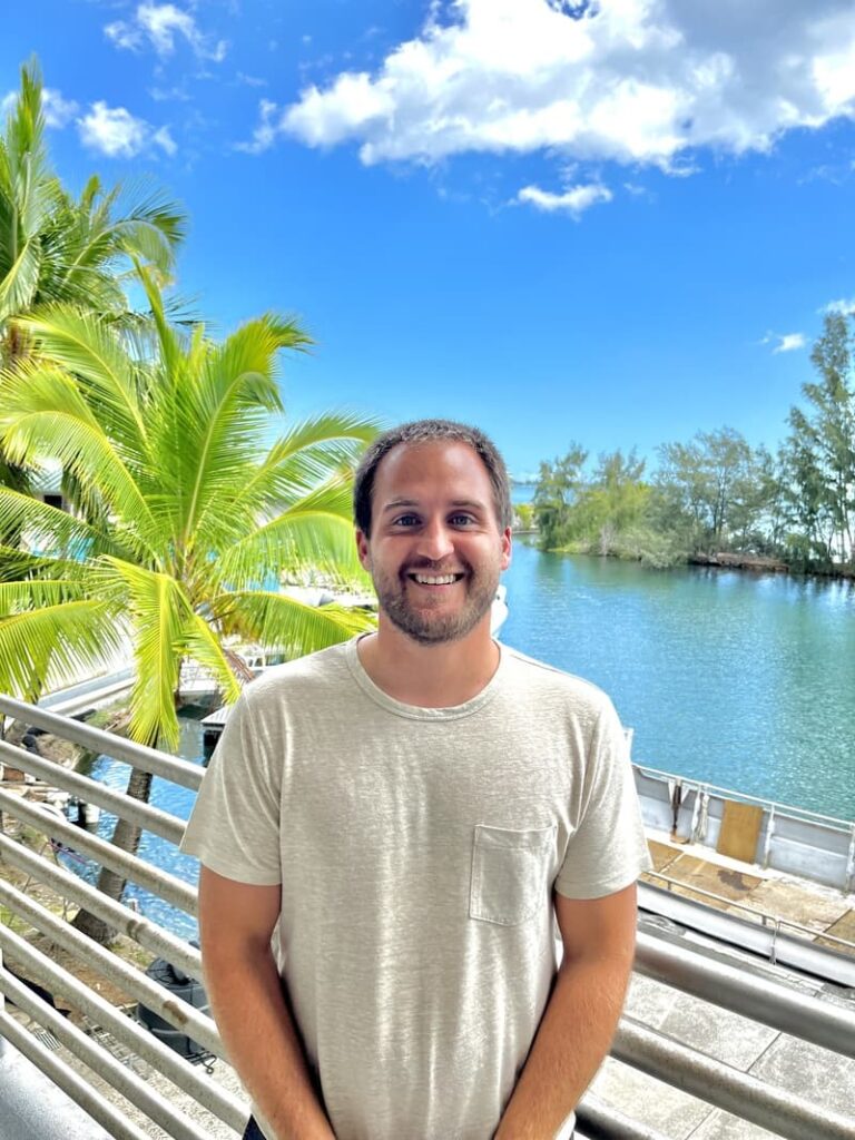 Smiling man in light t-shirt stands by railing with palm trees and water under a bright blue sky.