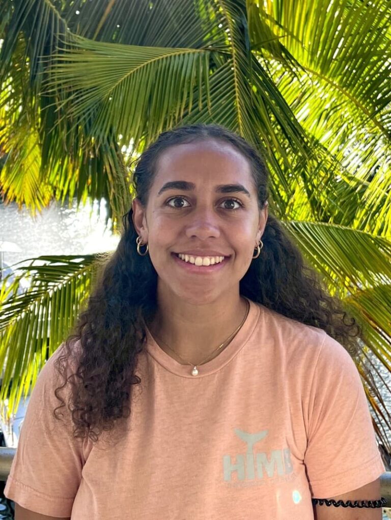 Smiling woman with curly hair in a pink shirt, standing in front of green palm leaves.