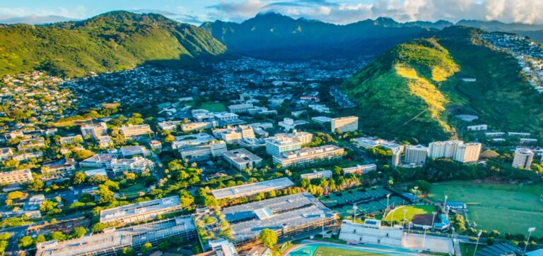 Aerial view of a city campus with green mountains and a stadium in the background, under a partly cloudy sky.